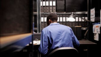 African american private investigator checking crime scene report, analyzing case evidence late at night in arhive room. Police officer in formal suit discovery confidential criminology files. Foto: Magnify.com