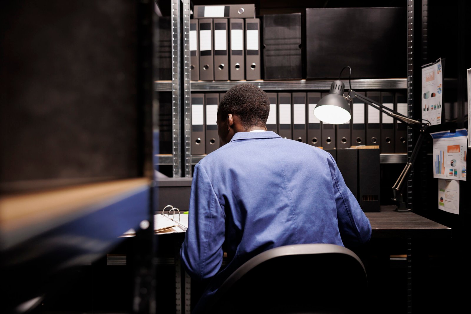 African american private investigator checking crime scene report, analyzing case evidence late at night in arhive room. Police officer in formal suit discovery confidential criminology files. Foto: Magnify.com